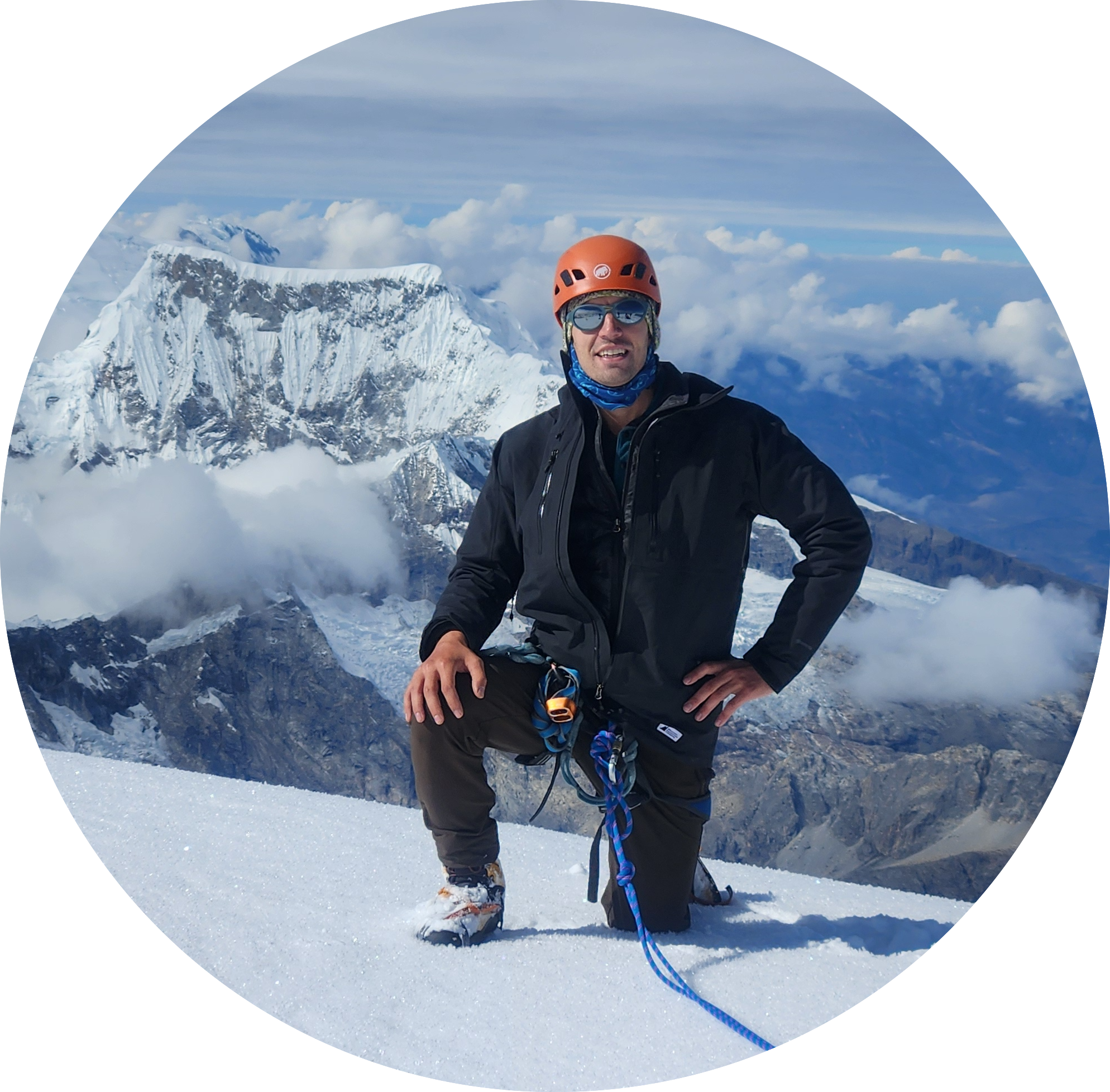 Duncan Steinke in mountaineering gear on a snow-covered summit, with glaciated peaks and cloud below.